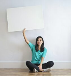 Cheerful Asian woman sitting cross legged on floor against white wall in empty apartment and showing white blank banner